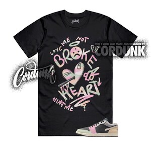 May include: Black t-shirt with a graphic design featuring the words "LOVE ME NOT BROKE HEART HURT ME" in a graffiti style. The design includes a broken heart with a knife. A pair of sneakers with black, pink, and beige accents are at the bottom.
