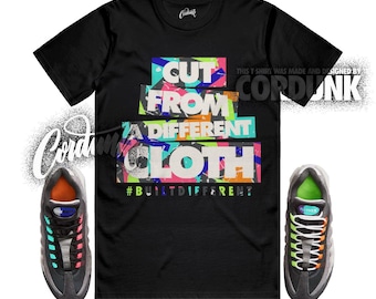 Peut inclure: T-shirt noir avec le texte "CUT FROM A DIFFERENT CLOTH" en lettres colorées. Le t-shirt comporte également le hashtag "#BUILTDIFFERENT". Deux baskets noires et grises avec des accents colorés sont de chaque côté du t-shirt.