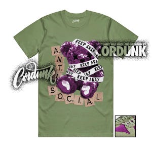 May include: Olive green t-shirt featuring a purple teddy bear graphic wrapped with white tape that reads "KEEP AWAY." The bear is surrounded by Scrabble tiles spelling "ANTI SOCIAL." The shirt also has the Cordunk logo.