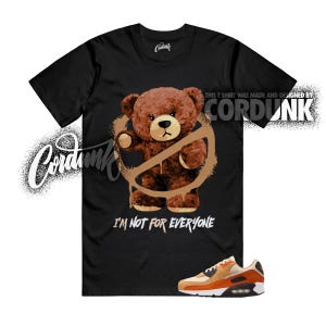 May include: Black t-shirt featuring a brown teddy bear with a graffiti-style prohibition symbol. The text "I'M NOT FOR EVERYONE" is below the bear. A pair of orange and tan sneakers is at the bottom.