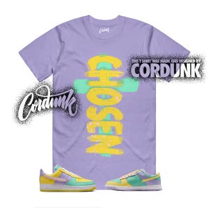 May include: A lilac t-shirt with the word "CHOSEN" in yellow and teal brushstrokes. The shirt is paired with yellow, teal, and purple sneakers. The text "THIS T SHIRT WAS MADE AND DESIGNED BY CORDUNK" is printed on the shirt.