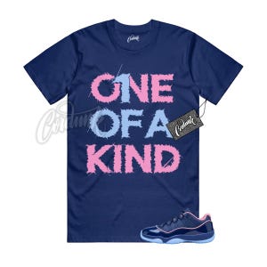 May include: A navy blue short-sleeve t-shirt featuring the text "ONE OF A KIND" in a distressed, jagged font with alternating light blue and pink letters. A black rectangular hang tag with the brand name "Cordunk" is attached to the sleeve.