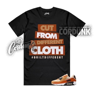 May include: Black t-shirt with the text "CUT FROM DIFFERENT CLOTH" in white and brown block letters. The phrase "#BUILTDIFFERENT" is below the text. A pair of orange and tan sneakers is at the bottom. The brand name "Cordunk" is in the upper right.