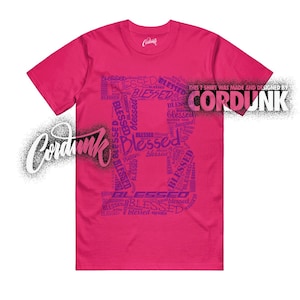 May include: A magenta t-shirt with the word "Blessed" in a stylized design. The word "Blessed" is repeated in a pattern to form the letter "B" on the front of the shirt. The text "THIS T SHIRT WAS MADE AND DESIGNED BY CORDUNK" is printed on the upper right.