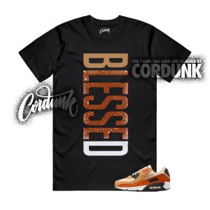 May include: Black t-shirt with the word "BLESSED" in a vertical, textured, orange and white design. The shirt also features a pair of orange and white sneakers. The text "THIS T SHIRT WAS MADE AND DESIGNED BY CORDUNK" is also visible.