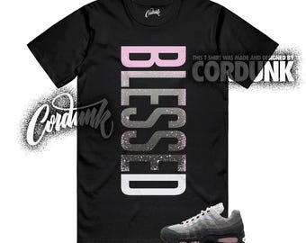 May include: Black t-shirt with the word "BLESSED" in a vertical design, featuring pink, gray, and white colors. The shirt is paired with gray and black sneakers with pink accents. The text "THIS T SHIRT WAS MADE AND DESIGNED BY CORDUNK" is also visible.
