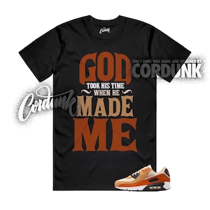 May include: Black t-shirt with the text "GOD TOOK HIS TIME WHEN HE MADE ME" in brown and tan. A pair of orange and tan sneakers is at the bottom. The shirt is designed by Cordunk.