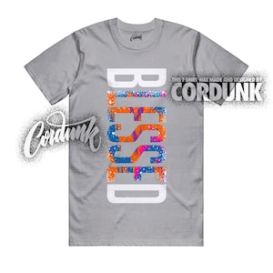 May include: A gray t-shirt with the word "BLESSED" in large, colorful letters down the front. The word is white with orange, blue, and pink accents. The brand name "CORDUNK" is also visible.