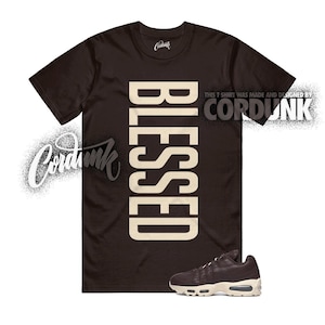May include: A dark brown t-shirt with the word "BLESSED" in large, cream-colored letters. The shirt also features the text "THIS T SHIRT WAS MADE AND DESIGNED BY CORDUNK". A pair of brown and cream sneakers is at the bottom.