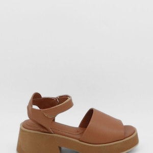 May include: A brown leather sandal with a platform sole. The sandal has a wide strap over the toes and an ankle strap with a buckle. The platform sole is approximately 5 cm high.