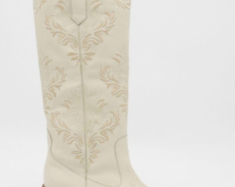 Peut inclure: Botte de cowboy crème avec broderies florales complexes. La botte haute présente un bout pointu, un talon épais et une tirette. La broderie est d'une teinte plus claire, créant un contraste subtil. La botte est en cuir.