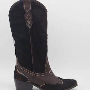 May include: A tall, brown and black cowboy boot with a pointed toe and a block heel. The boot features a dark brown leather vamp and a black, textured shaft. White stitching accents the design. The boot is set against a plain white background.