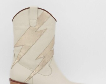 Peut inclure: Botte de cow-boy crème avec un motif d'éclair doré. La botte a un bout pointu, un talon en bois empilé et une bordure marron. Le motif d'éclair est sur le côté de la botte. Hauteur du talon : environ 5 cm.