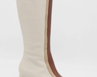 May include: Cream-colored knee-high boots with a brown leather accent on the side and toe. The boots have a block heel and a small gold-colored button near the top. The design includes a tan-colored trim.
