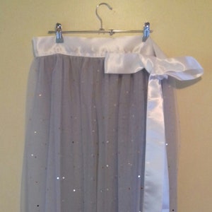 Può includere: Una gonna di tulle bianca con una cintura di raso bianco e un fiocco di raso bianco. La gonna è decorata con piccole paillettes argentate.