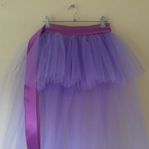 Può includere: Una gonna di tulle lavanda con un nastro di raso viola. La gonna è a strati e ha un orlo arricciato.