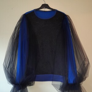 Può includere: Una camicia a maniche lunghe blu e nera con maniche in tulle nero trasparente. Le maniche sono arricciate alle spalle e ai polsi, creando un look voluminoso.