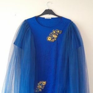 Può includere: Una t-shirt blu reale con un'sovrapposizione di tulle blu trasparente. La t-shirt ha un collo rotondo e maniche corte. La sovrapposizione di tulle si estende oltre l'orlo della t-shirt e ha un aspetto drappeggiato. La t-shirt è decorata con un motivo floreale fatto di perline e paillettes.