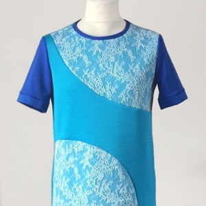 Può includere: Una maglietta blu con maniche corte e scollo tondo. La maglietta presenta un pannello turchese con dettagli in pizzo bianco. Le maniche e lo scollo sono di una tonalità di blu più scura, creando un design a contrasto.