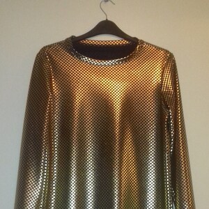 Può includere: Una camicia dorata a maniche lunghe con un motivo a quadretti strutturato. La camicia è appesa a una gruccia nera contro un muro bianco. Il tessuto ha un aspetto lucido e metallico, che riflette la luce.