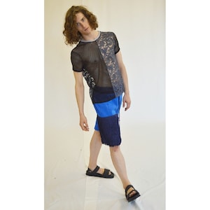 Può includere: Una persona che indossa una camicia di rete e pizzo nera e grigia con pantaloncini blu e neri. La persona indossa sandali.