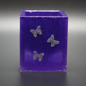 May include: A purple square-shaped container with three silver butterfly decorations.