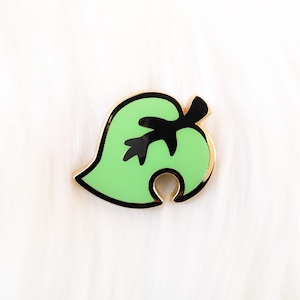 May include: A green leaf-shaped enamel pin with a black outline and a black stem. The pin is a popular symbol from the video game Animal Crossing.