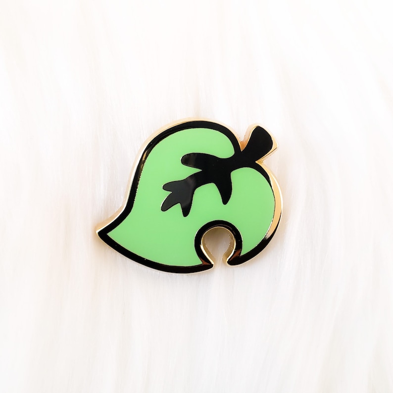 May include: A green leaf-shaped enamel pin with a black outline and a black stem. The pin is a popular symbol from the video game Animal Crossing.