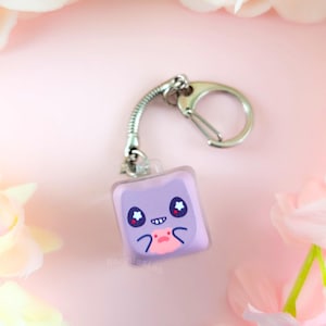 May include: A square-shaped purple keychain featuring a cartoon character with large star-filled eyes, a small smiling mouth, and a pink belly patch. The charm is attached to a silver-toned metal split ring by a short, flexible coiled metal spring.