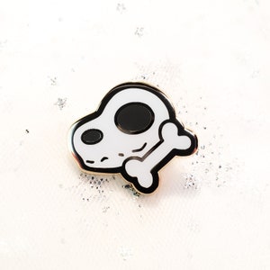 May include: A white enamel pin with a black outline of a cartoon dog skull holding a bone. The pin has a gold finish.