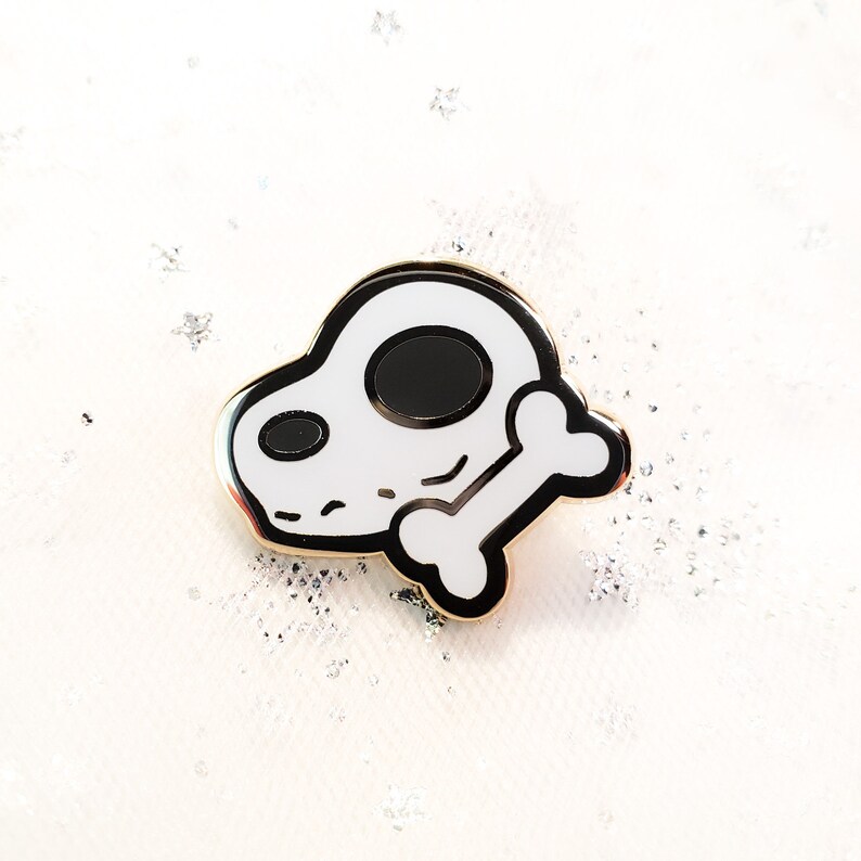 May include: A white enamel pin with a black outline of a cartoon dog skull holding a bone. The pin has a gold finish.