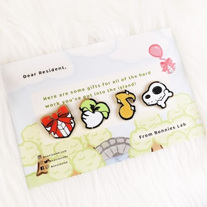 May include: A set of five gold enamel pins featuring a red gift box with a bow, a green apple, a yellow musical note, a white skull with a bone, and a pink balloon with a ribbon. The pins are on a white card with the text "Dear Resident, Here are some gifts for all of the hard work you've put into the island! From Bonnies Lab" printed on it.