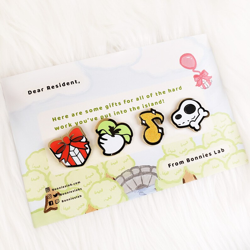 May include: A set of five gold enamel pins featuring a red gift box with a bow, a green apple, a yellow musical note, a white skull with a bone, and a pink balloon with a ribbon. The pins are on a white card with the text "Dear Resident, Here are some gifts for all of the hard work you've put into the island! From Bonnies Lab" printed on it.