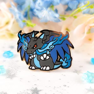 May include: A detailed enamel pin featuring a stylized dragon with black, gray, and blue coloring. The dragon has a fierce expression and is surrounded by blue flame-like accents. The pin has a gold-colored border and is set against a soft, blurred background.