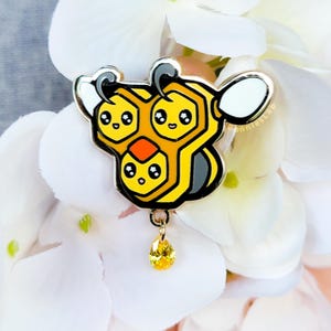 May include: A gold-colored enamel pin featuring a honeycomb design with three smiling bee faces. The pin has black, yellow, and white accents, and a dangling yellow teardrop gem. The pin is set against a backdrop of white flowers.