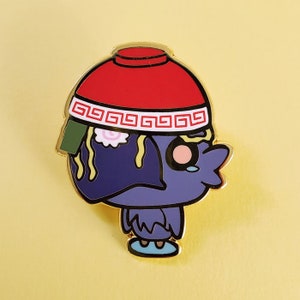 May include: A gold-colored enamel pin featuring a cartoon bird wearing a red and white bowl on its head. The bowl has a gold rim and a pattern of squares. The bird has a blue body, pink cheeks, and yellow noodles coming out of the bowl.