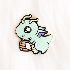 May include: A light green dragon with purple wings carrying a small brown bucket. The dragon has a large black eye with a white star in the center. The dragon is a cartoon style enamel pin.