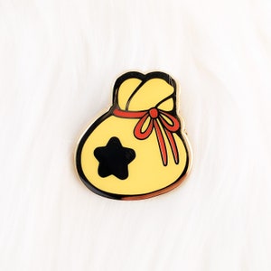 May include: A yellow and black enamel pin shaped like a money bag with a red ribbon and a star on the front.