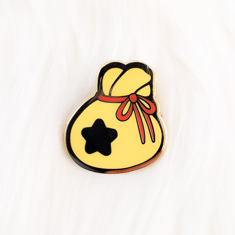 May include: A yellow and black enamel pin shaped like a money bag with a red ribbon and a star on the front.