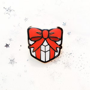May include: A gold-colored enamel pin shaped like a gift box with a red bow. The gift box is white with red stripes.
