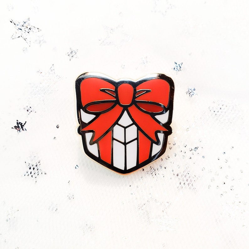 May include: A gold-colored enamel pin shaped like a gift box with a red bow. The gift box is white with red stripes.