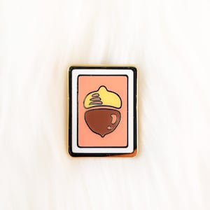 May include: Gold-toned enamel pin with a white border, featuring a cartoon acorn with a brown cap and a yellow top on a pink background.
