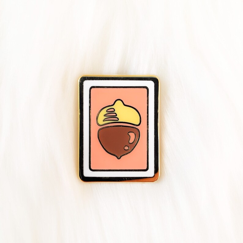 May include: Gold-toned enamel pin with a white border, featuring a cartoon acorn with a brown cap and a yellow top on a pink background.