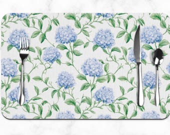 May include: Rectangular placemat featuring a watercolor floral design of blue hydrangeas and green leaves on a white background. A fork, knife, and spoon are placed on the placemat. The placemat is designed for dining and kitchen use.