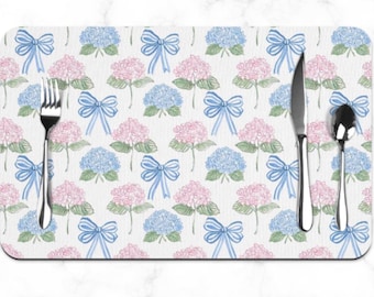 May include: Rectangular placemat with a floral pattern of pink and blue hydrangeas and blue bows. A fork, knife, and spoon are placed on the placemat. The background is white.