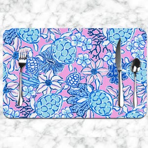May include: A rectangular placemat with a pink background and a pattern of blue and white flowers, sea turtles, and coral. A fork, knife, and spoon are placed on the placemat.