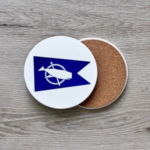 May include: A white ceramic coaster with a blue and white whale flag design. The coaster has a cork backing.