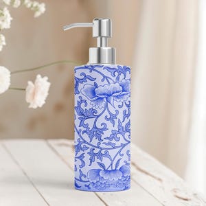 May include: A cylindrical soap dispenser featuring a light blue base with a dark blue floral pattern. The dispenser has a metallic silver pump mechanism at the top. The design consists of intricate, repeating botanical motifs of flowers and leaves.