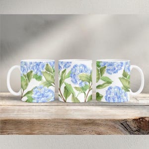 May include: Three white ceramic mugs with a blue hydrangea floral design. The mugs feature a repeating pattern of blue flowers and green leaves on a white background. The mugs are displayed on a wooden surface.