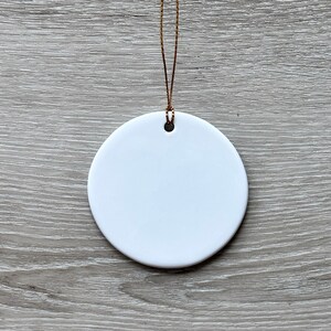 May include: A white ceramic ornament with a gold string attached. The ornament is round and has a hole in the top for hanging.